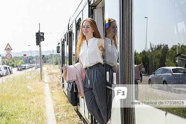 Lächelnde weibliche Fachkraft  die wegschaut  während sie mit einer Tasche vor einer Zugtür steht