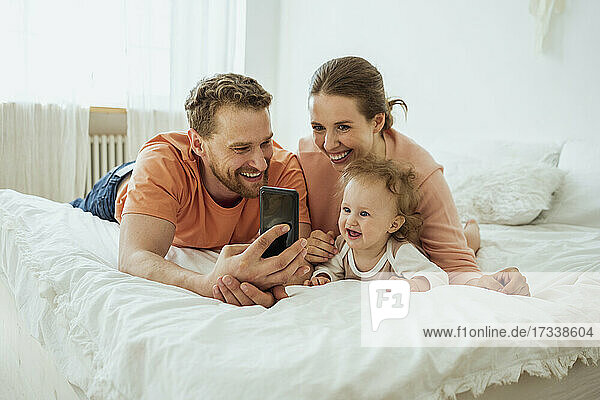 Lächelnder Mann zeigt Frau mit Tochter auf dem Bett zu Hause das Mobiltelefon