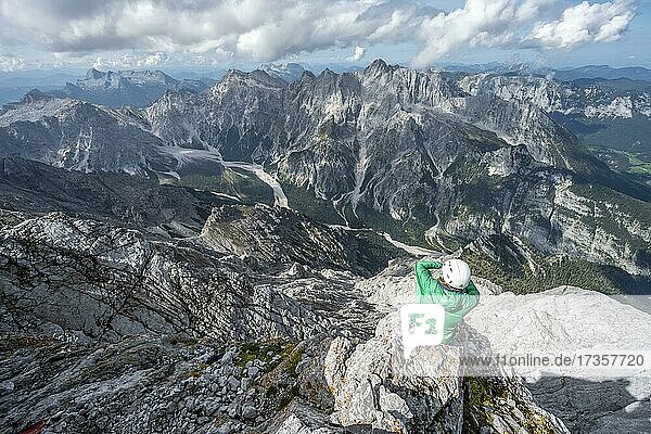 Wanderin mit Helm am Gipfel des Watzmann  Ausblick über Berge  Gebirgszug Hochkalterstock mit Blaueisspitze und Hochkalter  Wanderweg zum Watzmann  Watzmann-Überschreitung  Berchtesgaden  Bayern  Deutschland  Europa
