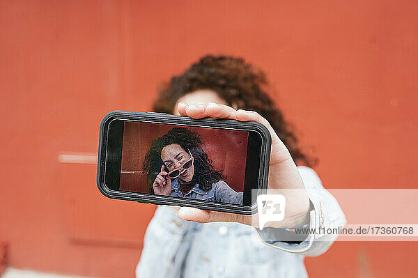 Junge Frau zwinkert  während sie ein Selfie mit ihrem Smartphone vor einer roten Wand macht