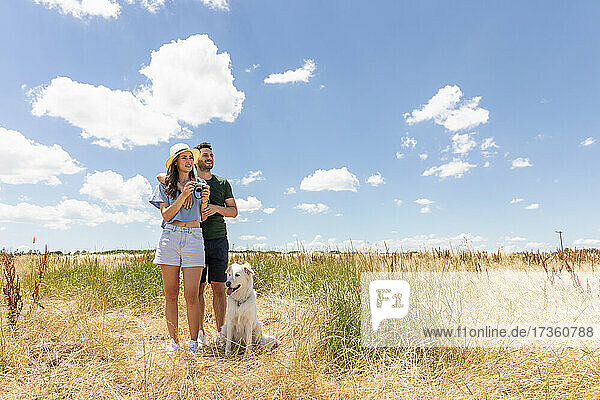 Woman holding camera while standing by boyfriend and dog at meadow