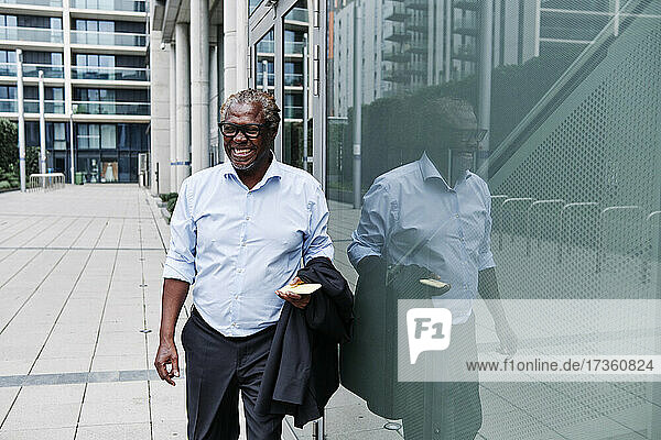 Happy businessman with smart phone and blazer walking by reflection on glass wall