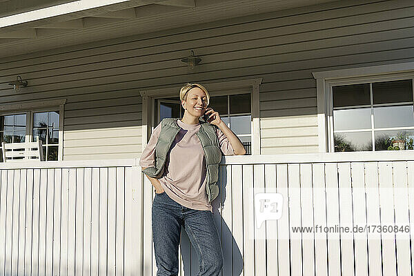 Frau spricht mit ihrem Smartphone und lehnt sich an ein Geländer vor dem Haus