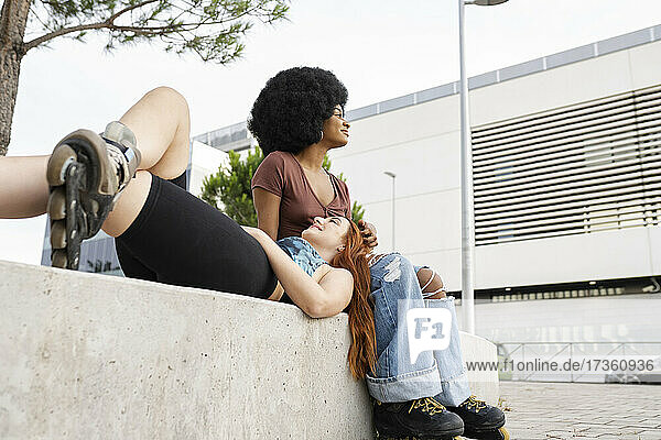 Afro woman looking away while female friend lying on lap in city