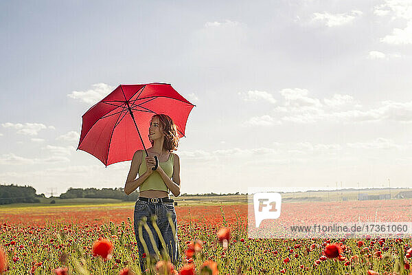 Teenage girl looking away while holding umbrella in poppy field