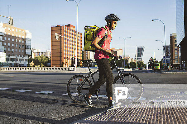 Zusteller  der eine Kiste hält  während er die Straße mit dem Fahrrad in der Stadt überquert