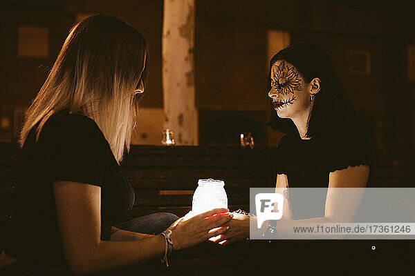 Freundinnen sitzen mit einem beleuchteten Gefäß auf einer Bank während Halloween