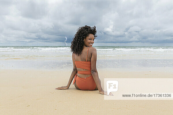 Lächelnde junge Frau  die über die Schulter zurückblickt  während sie am Strand sitzt