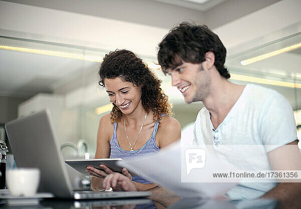 Lächelnder Mann  der einen Laptop benutzt  während seine Freundin ein Dokument in der Küche hält