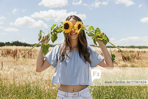 Woman covering eyes with sunflowers while standing at meadow