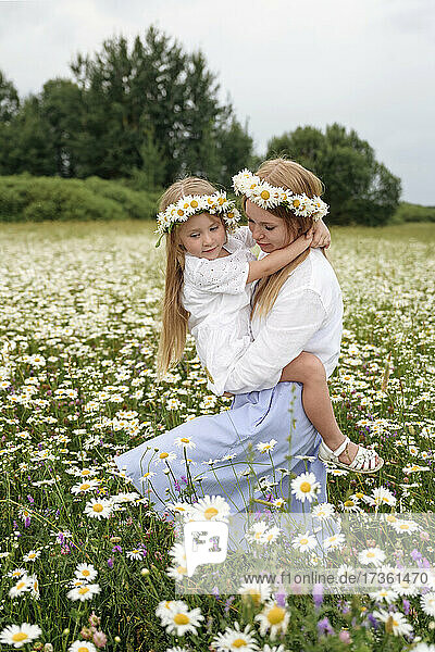 Woman with flower tiaras carrying daughter while walking at chamomile field
