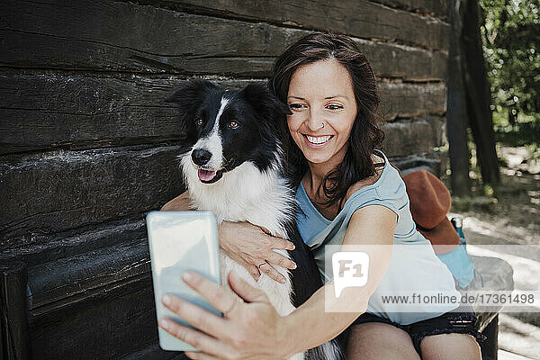 Lächelnde Frau nimmt Selfie mit Haustier Hund durch Smartphone  während außerhalb Hütte im Wald sitzen