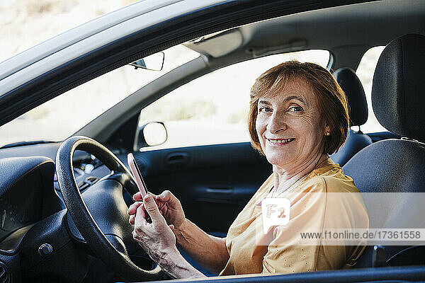 Lächelnde ältere Frau hält ihr Smartphone im Auto sitzend