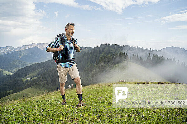 Lächelnder Mann mit Rucksack beim Wandern auf einer Wiese bei nebligem Wetter
