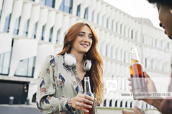 Smiling redhead woman holding beer bottle talking with female friend in city