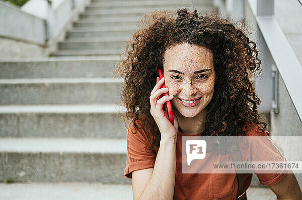 Smiling curly haired woman talking on mobile phone sitting at staircase