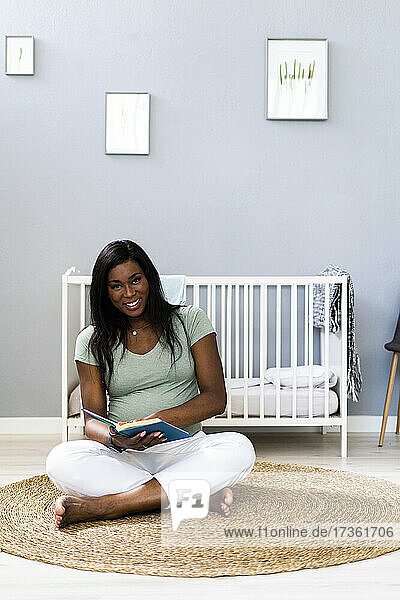 Smiling pregnant young woman sitting cross-legged with book in bedroom