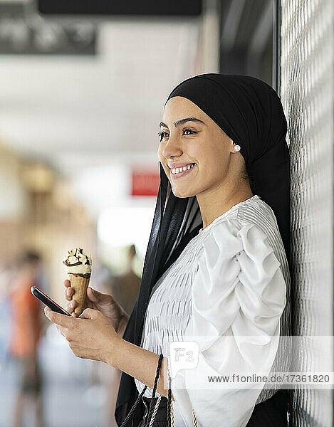 Lächelnde Frau mit Mobiltelefon  die ein Eis isst  während sie sich an die Wand lehnt