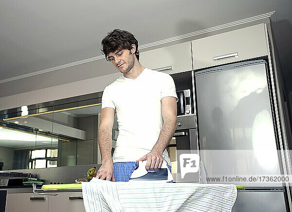 Junger Mann bügelt Hemd in der Küche zu Hause