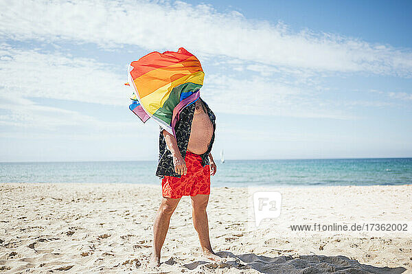 Schwuler Mann mit Regenbogenfahne im Gesicht am Strand im Urlaub stehend
