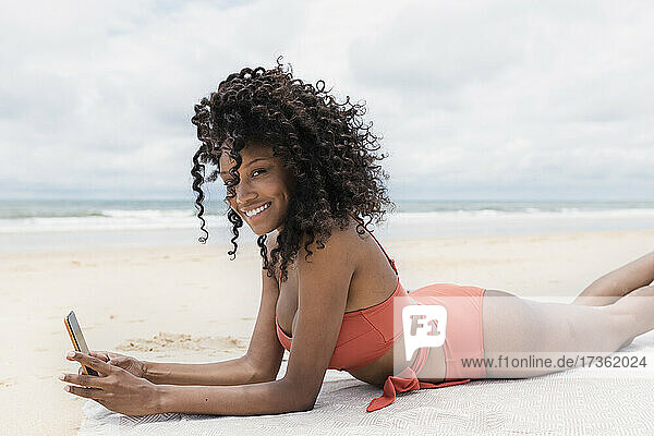 Lächelnde junge Frau mit Handy in der Hand am Strand liegend