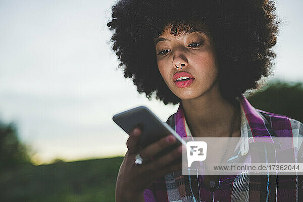 Porträt einer jungen Frau mit Afrofrisur  die in der Abenddämmerung im Freien ihr Smartphone benutzt