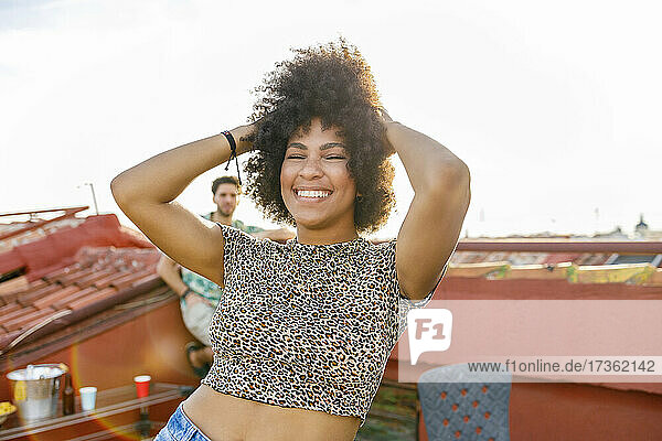 Cheerful Afro woman dancing with hands in hair on rooftop