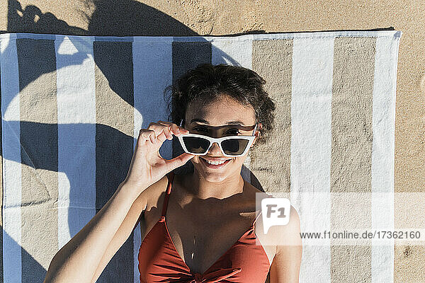 Lächelnde junge Frau mit Sonnenbrille auf einem Handtuch am Strand liegend