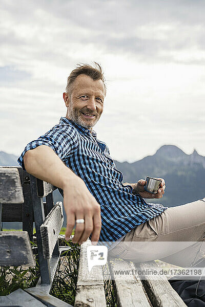 Smiling man holding coffee cup while sitting on bench