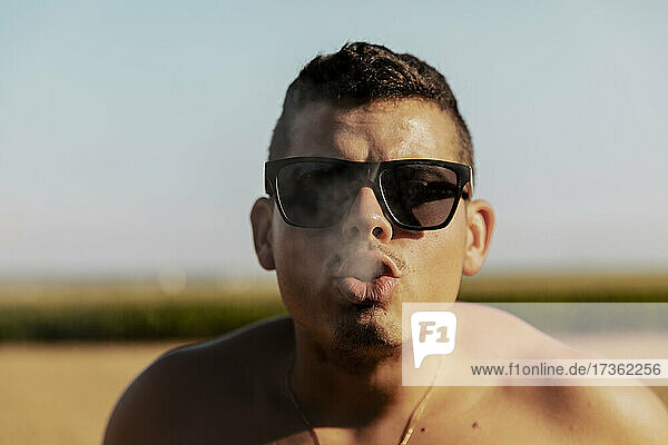 Man wearing sunglasses blowing smoke on sunny day