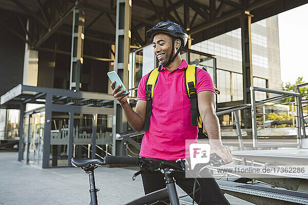 Lächelnder Zusteller  der eine SMS über sein Smartphone verschickt  während er mit seinem Fahrrad steht