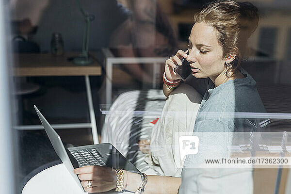 Businesswoman talking on mobile phone while using laptop seen from apartment window