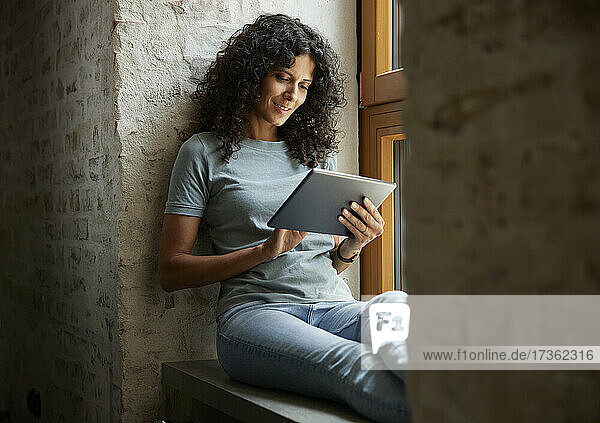 Businesswoman using digital tablet while sitting on window sill at workplace