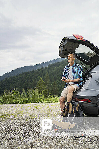 Männlicher Wanderer sitzt mit Handy im Kofferraum eines Autos