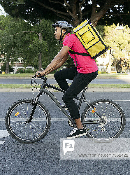 Männlicher Zusteller mit gelbem Rucksack fährt mit dem Fahrrad auf der Straße