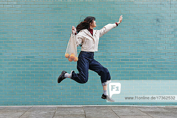 Excited young woman jumping while holding mesh bag by turquoise brick wall