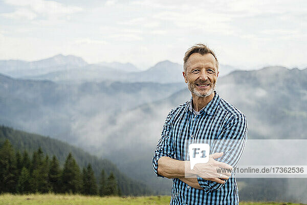 Lächelnder Mann mit verschränkten Armen auf einem Berg stehend