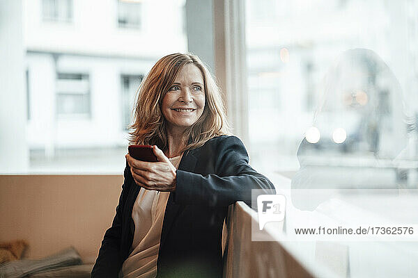 Lächelnde blonde weibliche Fachkraft  die ihr Smartphone hält und durch das Fenster schaut  während sie in einem Café sitzt