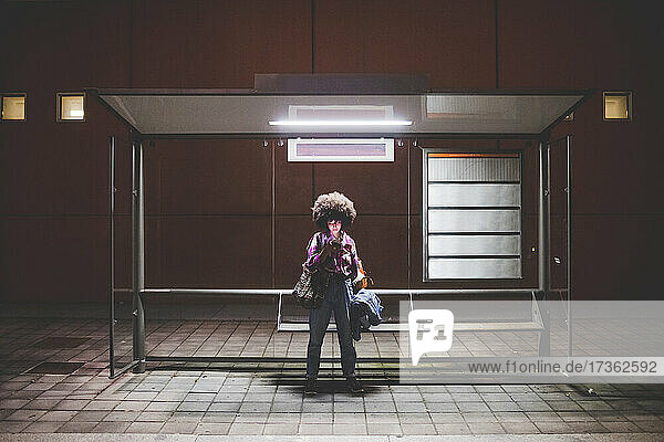 Young woman with afro hairdo using smartphone at bus stop in the city