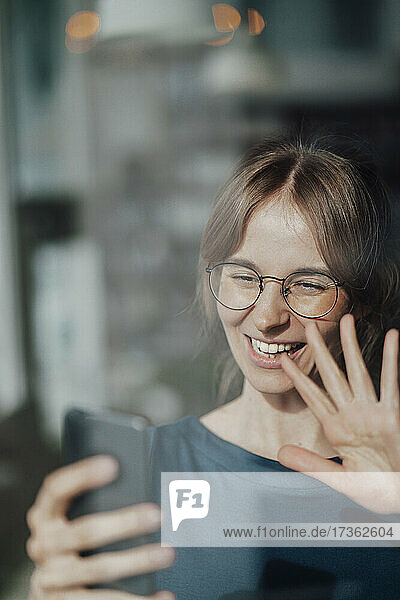 Glückliche Geschäftsfrau winkt mit der Hand  während sie einen Videoanruf über ein Smartphone tätigt