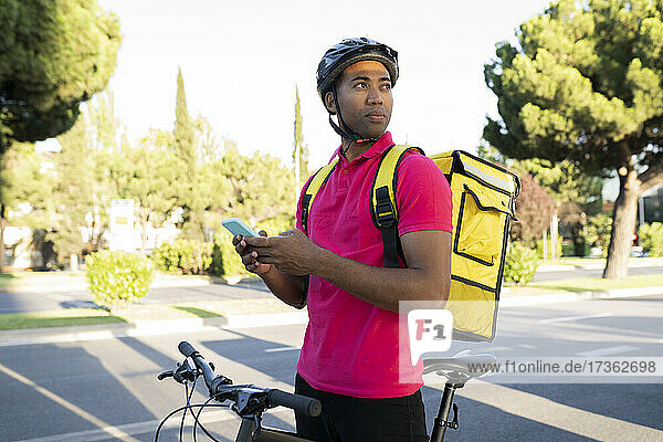 Delivery man holding smart phone looking away while standing with bicycle