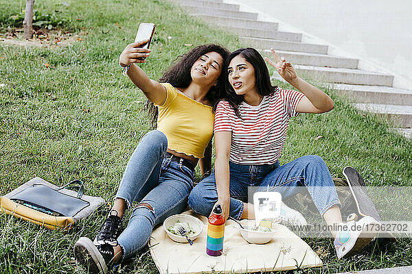 Junge Frau nimmt Selfie durch Handy mit weiblichen Freund gestikulieren Friedenszeichen beim Sitzen im Park