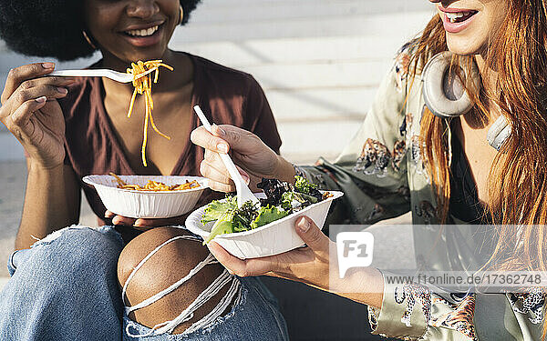 Lächelnde junge Freundinnen essen Salat und Spaghetti an einem sonnigen Tag
