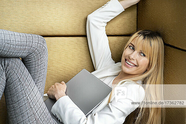 Lächelnde blonde Geschäftsfrau hält ein digitales Tablet  während sie sich auf dem Sitz zurücklehnt