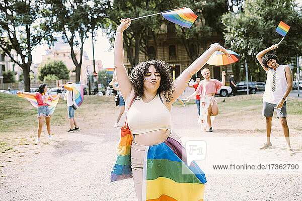 Fröhliche Frau hält Regenbogenfahnen mit Freunden im Hintergrund im Park