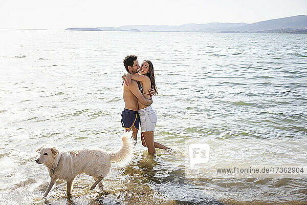 Smiling couple embracing while standing by dog in lake