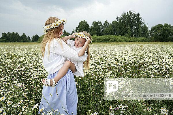 Blonde Frau trägt ein Mädchen  das Blumen trägt  während sie auf einem Blumenfeld steht
