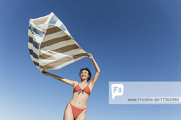 Lächelnde junge Frau winkt mit Handtuch vor blauem Himmel