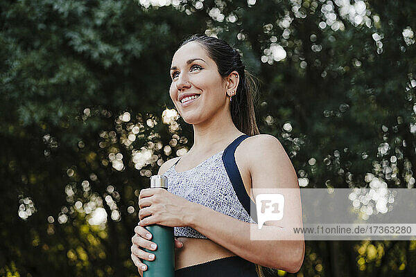 Lächelnde Sportlerin schaut weg  während sie eine Wasserflasche im Wald hält