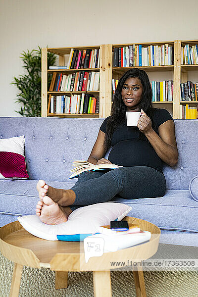 Nachdenkliche schwangere Frau hält eine Kaffeetasse  während sie zu Hause auf dem Sofa sitzt